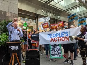 Sen. Bernie Sanders speaks in front of a crowd of ten people carrying signs that read "People over profits"