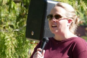 A woman in a burgundy sweater speaks into a microphone outside.
