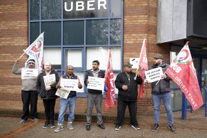 A group of men stand in front of a building with "uber" on it holding signs about worker rights