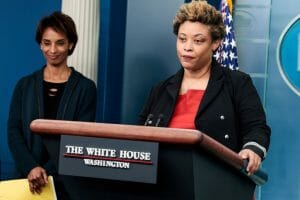 A woman stands at a podium with a sign that reads: The White House