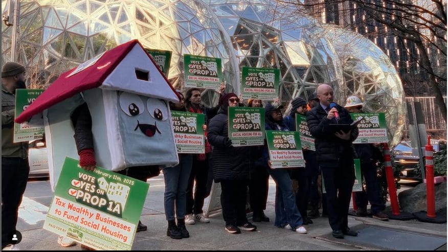 A group of people at a rally with signs saying ''tax the wealthy to pay for public housing''